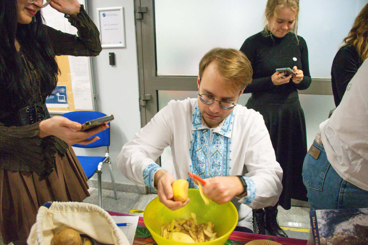 Grupa osób w pomieszczeniu, na pierwszym planie mężczyzna siedzący przy stole obiera ziemniaka nad dużą zieloną miską z obierkami. Ma jasne włosy, okulary i białą koszulę z niebieskim wzorem na przodzie i mankietach. Na stole widać kolorowy obrus oraz z lewej strony koszyk lub worek z ziemniakami. W tle stoją dwie kobiety ubrane na ciemno, trzymają telefony; po prawej w kadrze częściowo widać kolejną osobę w jasnej koszuli i dżinsach. W pomieszczeniu są krzesła i drzwi oraz tablica lub panel na ścianie.