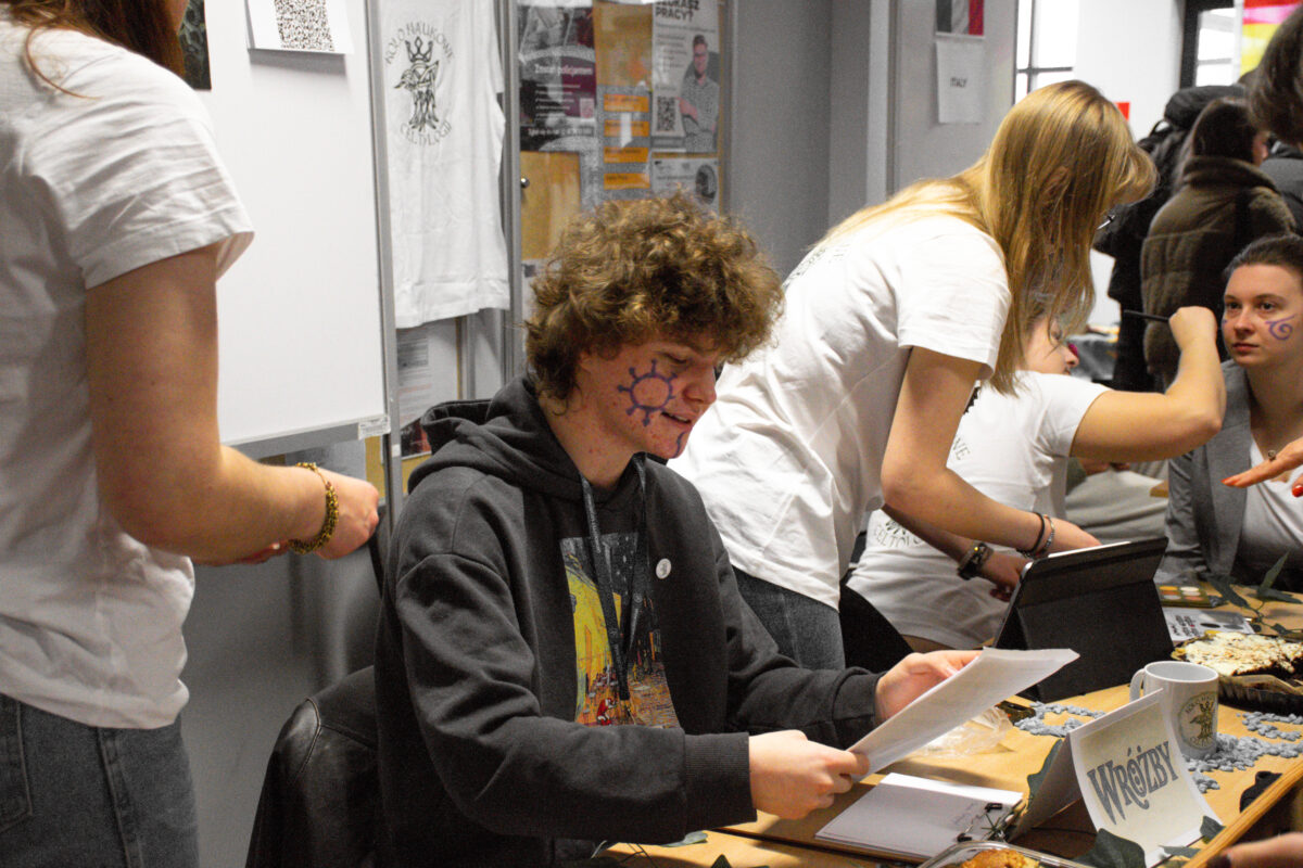 Scena w pomieszczeniu z kilkoma osobami przy stole, przypominająca stoisko informacyjne lub punkt rekrutacyjny. W centrum siedzi młoda osoba o jasnej cerze i kręconych brązowych włosach, w ciemnej bluzie z kapturem, trzyma kartkę i czyta. Na lewym policzku ma namalowany fioletowy symbol przypominający słońce. Po prawej stronie dwie osoby w białych koszulkach pochylają się nad stołem; jedna z nich wygląda na to, że maluje lub rysuje coś na twarzy innej osoby. Na stole leżą dokumenty, segregator, laptop, kubek oraz tabliczka z napisem „WRÓŻBY”. W tle widać tablicę i ścianę z plakatami oraz koszulkę z nadrukiem zawieszoną na ścianie.