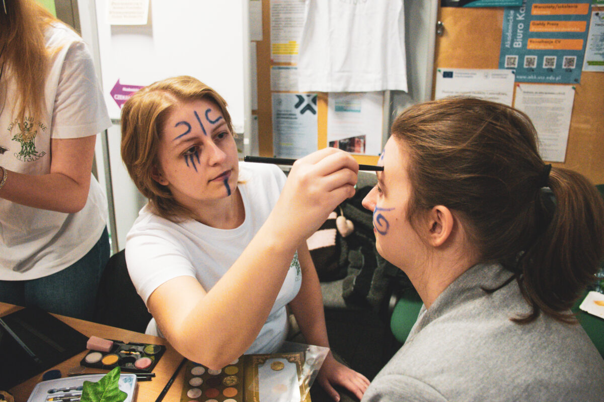 Dwie młode kobiety w pomieszczeniu, jedna maluje drugiej twarz pędzlem. Kobieta po lewej ma jasne, krótko ścięte blond włosy i białą koszulkę; na czole ma znaczki, a na policzku i pod okiem ciemnoniebieskie, pionowe kreski. Kobieta po prawej ma brązowe włosy związane w kucyk, jasną cerę i szarą bluzę; na policzku ma namalowany niebieski wzór spirali. Na stole widać otwartą paletę farbek/paintów do makijażu i pędzle. W tle są tablice i kartki z ogłoszeniami oraz biała tkanina zawieszona na ścianie.