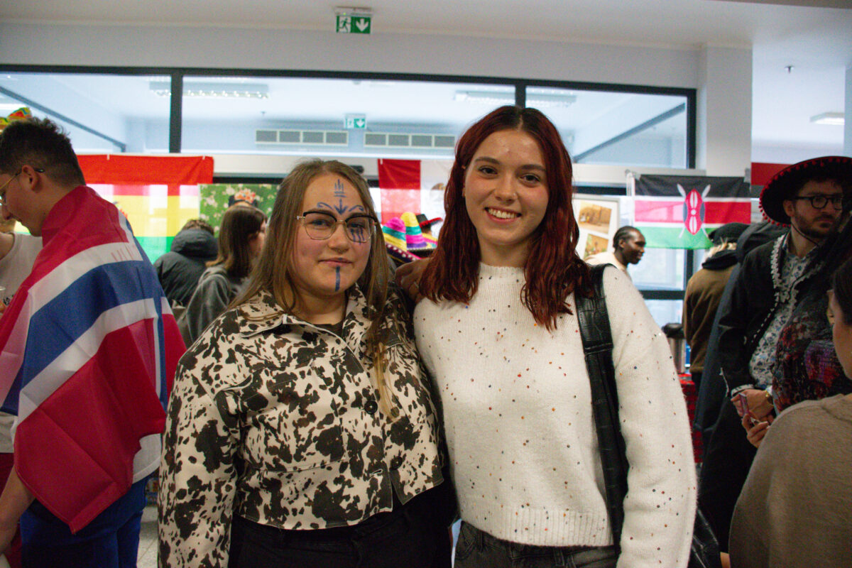 Dwie młode kobiety stojące blisko siebie w zatłoczonym pomieszczeniu, na tle wiszących flag. To wygląda na wydarzenie sali z dużymi oknami i jasnym oświetleniem. Kobieta po lewej ma jasną cerę, proste jasnobrązowe włosy do ramion i okulary. Na twarzy ma namalowane niebieskie linie: pionową na czole i nosie oraz pionową na brodzie. Ma na sobie jasną kurtkę w czarno-biały wzór i ciemne spodnie. Kobieta po prawej ma jasną cerę i falowane, ciemnorude włosy do ramion. Uśmiecha się, ma na sobie biały sweter z drobnymi kolorowymi kropkami oraz czarny pasek od torby na ramieniu. W tle widać kilka osób i różne flagi, m.in. po lewej flagę Wielkiej Brytanii na czyichś plecach, a po prawej flagę Kenii. Nad oknami są zielone znaki wyjścia ewakuacyjnego.
