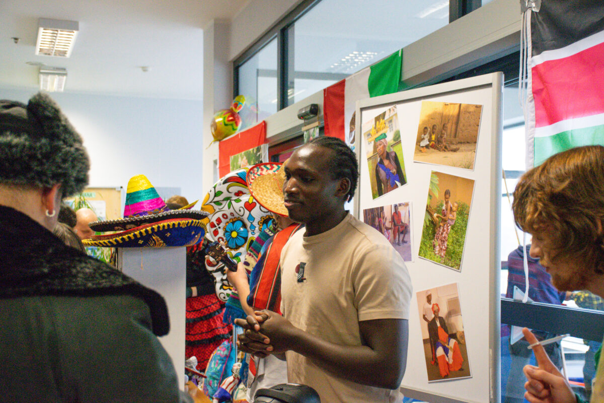 Grupa osób w pomieszczeniu, stojąca przy tablicy z przypiętymi zdjęciami i otoczona kolorowymi dekoracjami oraz flagami. W centrum stoi czarnoskóry mężczyzna z włosami związanymi z tyłu, w jasnobeżowej koszulce, z małym mikrofonem przypiętym na piersi; ma złożone dłonie i jest zwrócony w stronę innych osób, jak podczas rozmowy lub prezentacji. Po lewej stronie na podwyższeniu leży kolorowe sombrero; obok widać duży, wzorzysty, okrągły kapelusz oraz inne elementy w jaskrawych barwach. Po prawej stronie stoi biała tablica, do której przypięto kilka fotografii przedstawiających ludzi w różnych sytuacjach; nad tablicą i w tle wiszą flagi w barwach czerwono-biało-zielonych. W kadrze widać też fragmenty innych osób: z lewej plecy osoby w ciemnej kurtce i futrzanej czapce, a z prawej profil osoby o jasnobrązowych włosach.