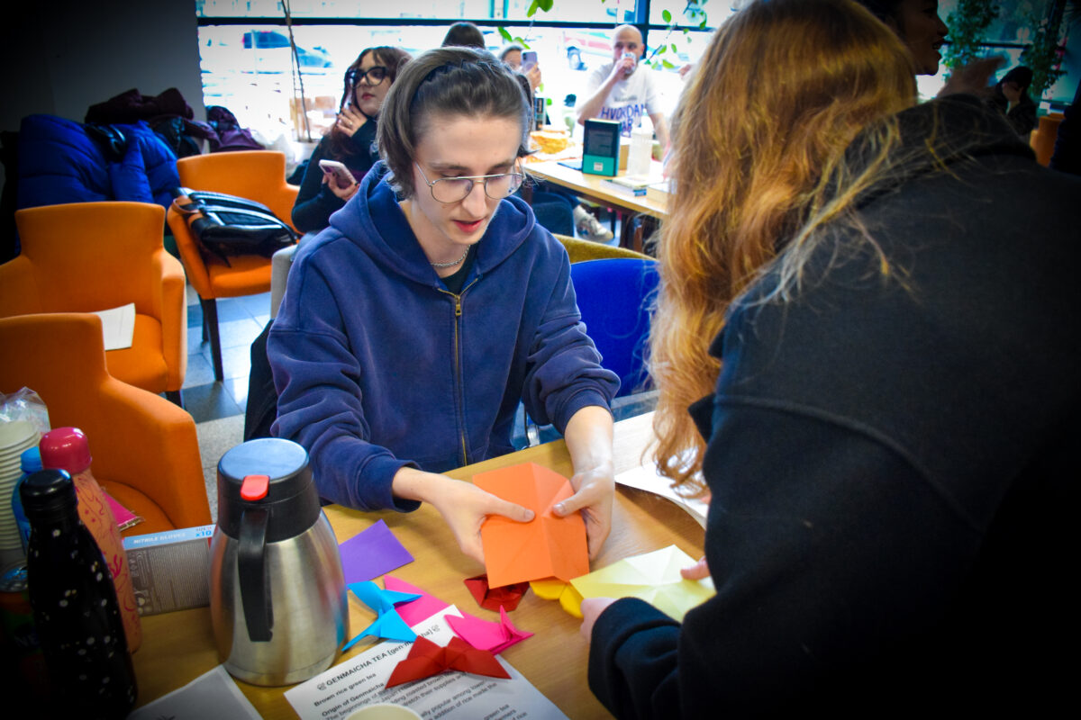 Ludzie przy stole w pomieszczeniu przypominającym kawiarnię lub świetlicę, z kolorowymi krzesłami w tle. Na pierwszym planie dwie osoby siedzą naprzeciwko siebie i składają papier (origami). Osoba po lewej ma jasną skórę, ciemne włosy związane z tyłu, okrągłe okulary i granatową bluzę z kapturem; trzyma w dłoniach duży pomarańczowy arkusz papieru. Osoba po prawej (widoczna od tyłu/boku) ma długie, falowane, rudawe włosy i ciemną bluzę; trzyma jasny, kremowo-żółty papier. Na stole leżą kolorowe, złożone papierowe kształty (różowe, niebieskie, czerwone i fioletowe) oraz kilka prostokątnych kartek papieru. Po lewej stronie stołu stoją butelki oraz metalowy dzbanek/termos. W tle widać inne osoby siedzące przy stolikach; jedna osoba trzyma telefon, a na dalszym planie przy stoliku stoi mały stojak/tabliczka. Na dole zdjęcia widać kartkę z częściowo czytelnym tekstem, ale jest zbyt mały i nieostry, by go wiernie przepisać.