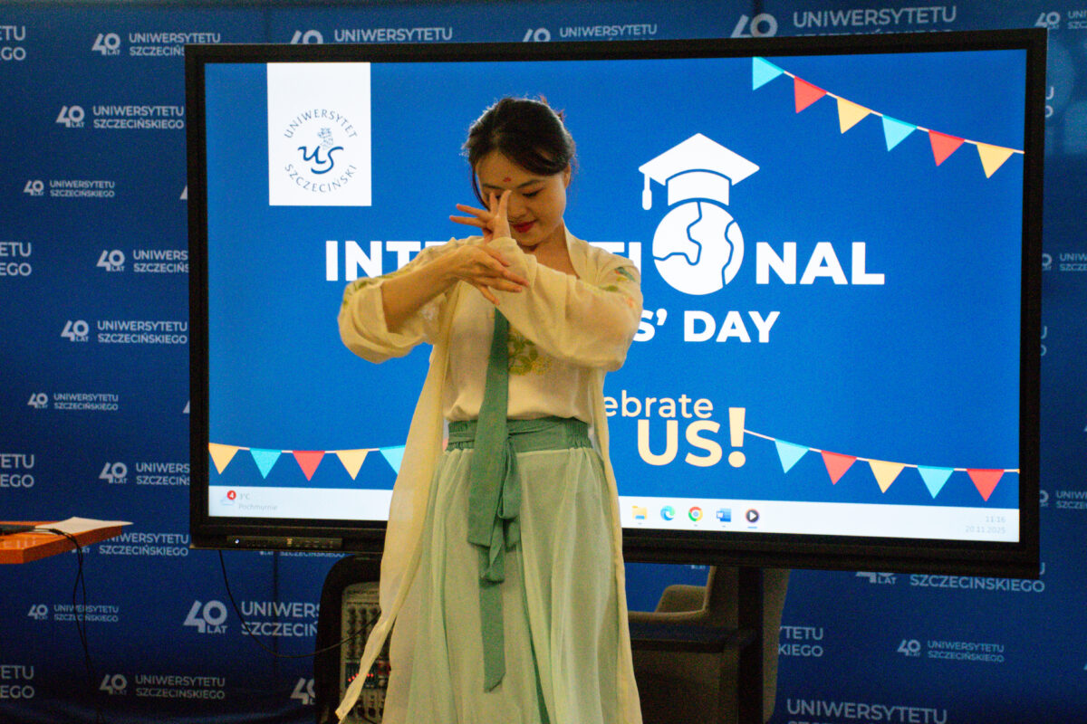 Młoda kobieta stojąca przed dużym ekranem z niebieskim slajdem wydarzenia. Ma ciemne włosy upięte do tyłu, jasną cerę (kobieta pochodzenia azjatyckiego) i jest ubrana w jasny, luźny strój z zielonkawą spódnicą/szerokimi spodniami oraz zielonym pasem lub szarfą w talii. Trzyma ręce uniesione przed twarzą w pozie przypominającej gest taneczny lub pantomimę. Na ekranie widać grafikę z napisem „INTERNATIONAL …’S DAY” oraz „Celebrate US!”, ikonę globusa w czapce absolwenta, a także małe, kolorowe chorągiewki w górnym prawym rogu. Po lewej stronie ekranu jest logo „Uniwersytet Szczeciński” z inicjałami „US”. Tło stanowi niebieska ścianka z powtarzającym się napisem „Uniwersytet Szczeciński” oraz „40 lat”. W dolnej części zdjęcia widać fragmenty sprzętu przy ekranie (podstawa/elementy stanowiska).