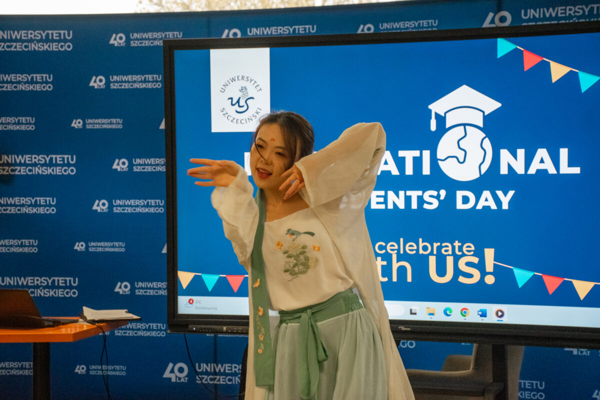 Scena wydarzenia na tle dużego ekranu z napisem „NATIONAL STUDENTS’ DAY” i „celebrate with us!”. W centrum stoi młoda kobieta w trakcie występu lub prezentacji, z uniesionymi rękami w pozie przypominającej taniec. Ma długie, ciemne włosy i lekki uśmiech. Ubrana jest w jasną, luźną bluzkę z długimi rękawami oraz zielonkawy strój z szerokim pasem i zwisającymi taśmami; na przodzie bluzki widać haftowany wzór roślinny. W tle widać logo „Uniwersytetu Szczecińskiego” oraz powtarzający się nadruk „Uniwersytetu Szczecińskiego” i „40 lat” na niebieskiej ściance. Po lewej stronie znajduje się stolik z laptopem.