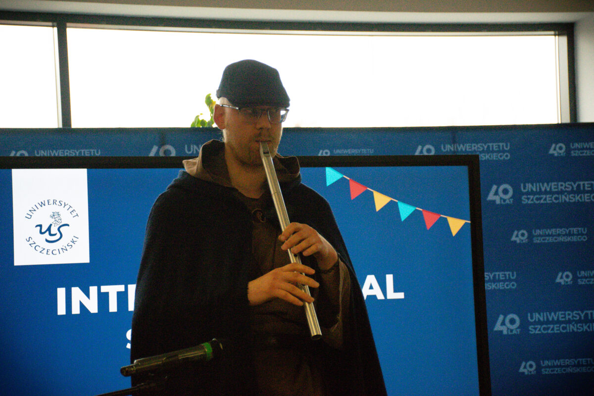 Mężczyzna w ciemnej czapce i okularach grający na metalowym flecie. Ma na sobie ciemną pelerynę lub płaszcz z kapturem. W tle są dwa duże ekrany lub tablice z niebieskim tłem. Po lewej widać logo i napis „UNIWERSYTET SZCZECIŃSKI” oraz stylizowane litery „US”. Na prawym ekranie jest girlanda z kolorowych trójkątnych chorągiewek. Po prawej stronie tła powtarza się napis „UNIWERSYTETU SZCZECIŃSKIEGO” oraz „40 LAT”. Na dole po lewej widoczny jest fragment mikrofonu na statywie.