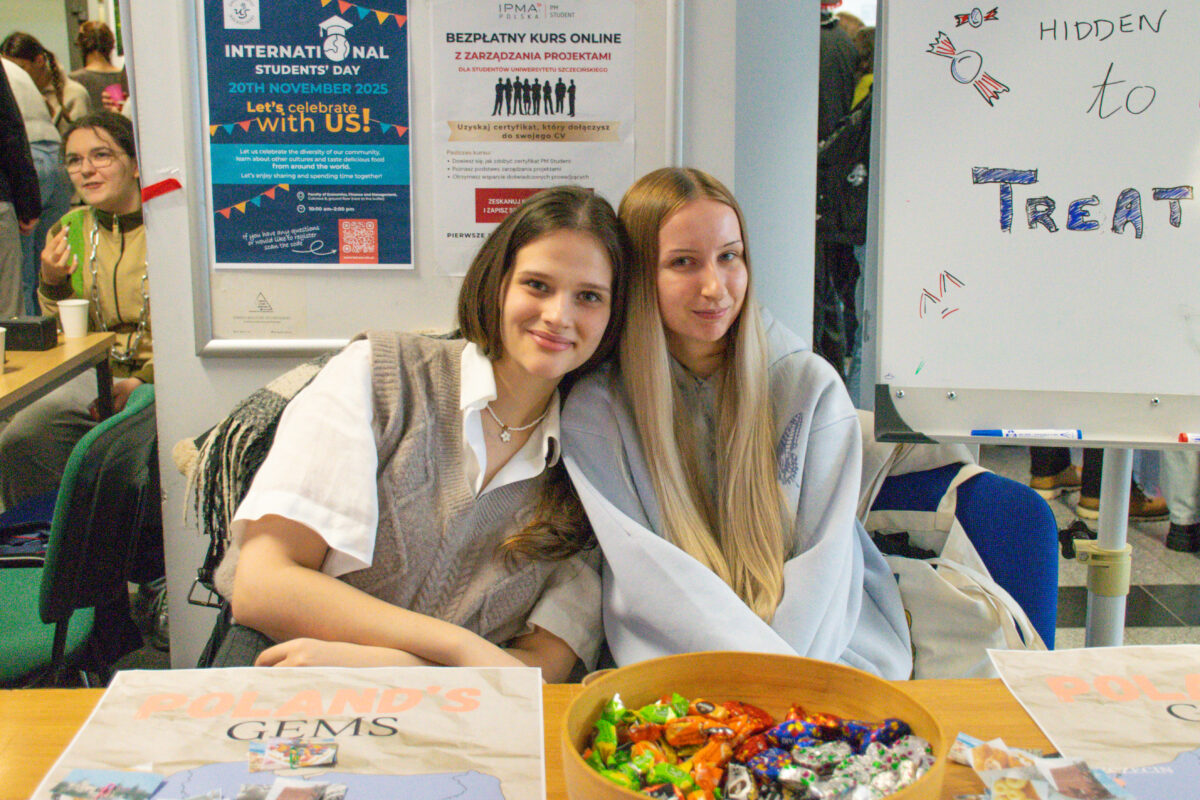 Dwie młode kobiety siedzące blisko siebie przy stole, uśmiechające się do aparatu. Obie mają jasną karnację. Kobieta po lewej ma ciemnobrązowe, proste włosy do ramion i ma na sobie białą koszulę oraz szarą, dzianinową kamizelkę; na szyi ma naszyjnik. Kobieta po prawej ma bardzo długie, proste, blond włosy i ma na sobie jasnoszarą bluzę. Na stole stoi duża, okrągła miska wypełniona kolorowymi słodyczami w papierkach. Po obu stronach miski leżą kartki/plakaty; na lewym widać duży napis „POLAND’S GEMS”. W tle, na ścianie, wiszą dwa plakaty. Lewy ma nagłówek „INTERNATIONAL STUDENTS’ DAY”, datę „20TH NOVEMBER 2025” i tekst „Let’s celebrate with us!”. Prawy jest po polsku i ma nagłówek „BEZPŁATNY KURS ONLINE z zarządzania projektami”, a u góry widnieje „IPMA POLSKA” oraz „PM STUDENT”. Po prawej stronie stoi biała tablica z napisem „HIDDEN to TREAT” oraz rysunkami cukierków. W tle po lewej widać jeszcze jedną osobę przy innym stole i kubek jednorazowy. W tle, na ścianie, wiszą dwa plakaty. Lewy ma nagłówek „INTERNATIONAL STUDENTS’ DAY”, datę „20TH NOVEMBER 2025” i tekst „Let’s celebrate with us!”. Prawy jest po polsku i ma nagłówek „BEZPŁATNY KURS ONLINE z zarządzania projektami”, a u góry widnieje „IPMA POLSKA” oraz „PM STUDENT”. Po prawej stronie stoi biała tablica z napisem „HIDDEN to TREAT” oraz rysunkami cukierków. W tle po lewej widać jeszcze jedną osobę przy innym stole i kubek jednorazowy.