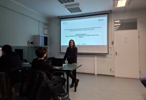 Sala wykładowa z projektorem i kilkoma osobami podczas zajęć. W centralnej części stoi kobieta o jasnej karnacji i ciemnych, rozpuszczonych włosach do ramion; ma okulary i ciemny żakiet, opiera się o blat zielonego stołu. Na stole leży zamknięty laptop i kilka kartek. Przed nią siedzi młody mężczyzna w ciemnej bluzie, bokiem do aparatu, przy biurku z otwartym laptopem; obok na oparciu krzesła wisi pikowana kurtka. Po lewej stronie widać jeszcze dwie osoby przy komputerach stacjonarnych, także w ciemnych ubraniach. Na ścianie za prowadzącą wisi rozwinięty biały ekran projekcyjny z wyświetlonym slajdem. Na górze slajdu widać logotypy, a tekst jest czytelny i brzmi: „Koło Naukowe Ekonomii Behawioralnej i Makroekonomii „IMPULS” Szkoła Giełdowa Fundacji GPW Uniwersytet Szczeciński Inwestycje giełdowe – kiedy na parkiecie tańczą fundamenty, technika i psychologia. Edycja 4.” Na dole slajdu znajdują się kolejne logotypy, w tym „GPW”. Pomieszczenie jest jasne, z białymi ścianami i sufitem podwieszanym z prostokątnymi lampami jarzeniowymi. Po prawej stronie kadru znajduje się białe skrzydło drzwiowe z klamką w kolorze złotym, obok gniazdko elektryczne z wpiętą wtyczką.