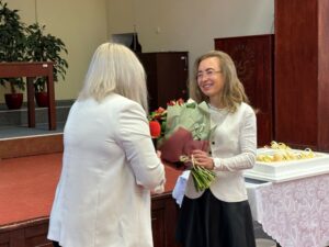 Sala Wydziału Ekonomii, Finansów i Zarządzania. Na zdjęciu 2 kobiety – prorektorka tyłem do kadru wręcza kwiaty dyrektorce Instytutu Ekonomii i Finansów (bokiem do kadru).