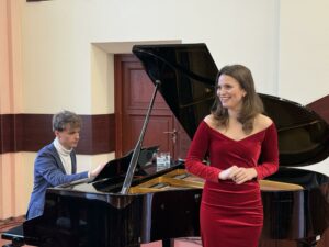 Sala Wydziału Ekonomii, Finansów i Zarządzania. Na pierwszym planie uśmiechnięta wokalistka (ujęcie do kolan). W tle czarny fortepian. Po lewej pianista.