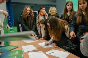 Grupa nastolatków stojących i pochylonych nad stołem, z dwiema dziewczynami podpisującymi formularze długopisami. Scena wygląda na wnętrze budynku, prawdopodobnie korytarz lub hol. Na stole leży kilka kartek z tabelami i miejscami na podpisy oraz parę długopisów. Po lewej stronie widać zielone teczki i zieloną torbę z białym napisem „ERASMUS!” oraz mniejszym tekstem poniżej (nieczytelnym). W tle stoi kilka osób w kurtkach i szalikach. Większość ma jasną skórę; dominują długie brązowe lub ciemnoblond włosy.