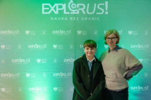 Zielone tło fotograficzne z dużym napisem „EXPLORUS!” i hasłem „NAUKA BEZ GRANIC” u góry. Na tle stoją dwie osoby, ujęte od pasa w górę, pozujące do zdjęcia. Osoba po lewej ma jasną skórę, krótkie brązowe włosy z grzywką i jest ubrana w ciemny płaszcz lub marynarkę, białą koszulę i krawat. Osoba po prawej ma jasną skórę, krótkie jasnobrązowe włosy, okulary i jest ubrana w jasny sweter z ciemniejszą koszulą pod spodem. Na tle wielokrotnie powtórzone są logotypy i napisy, m.in. „EXPLORUS! NAUKA BEZ GRANIC” oraz „Minister Edukacji”, a także małe znaki/embelmaty.