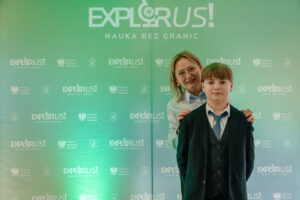 Zielone tło typu ścianka konferencyjna z dużym napisem „EXPLORUS!” u góry oraz hasłem „NAUKA BEZ GRANIC”. Na tle powtarzają się mniejsze logotypy „EXPLORUS! NAUKA BEZ GRANIC” oraz „Minister Edukacji”. Na pierwszym planie stoi chłopiec w ciemnym garniturze, białej koszuli i niebieskim krawacie. Za nim stoi kobieta w jasnej koszuli, w okularach, z krótkimi blond włosami; ma dłonie położone na jego ramionach. Oboje są zwróceni przodem do aparatu.