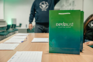Zielona papierowa torba z białym sznurkiem jako uchwytem, stojąca na drewnianym stole. Na torbie jest biały napis „EXPLORUS!” oraz mniejszy „NAUKA BEZ GRANIC”. Na stole leżą kartki z tabelami oraz długopisy. W tle w sali widoczna jest stojąca osoba w ciemnej bluzie z okrągłym białym nadrukiem na piersi oraz fragment siedzącej osoby po prawej stronie. Widać też krzesła i jasne ściany, jak w sali szkoleniowej lub klasie.
