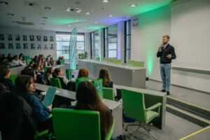 Sala wykładowa z publicznością siedzącą w zielonych fotelach, zwróconą w stronę mężczyzny stojącego z mikrofonem przy ścianie. Mężczyzna ma krótko przycięte włosy i krótki zarost, jasną karnację, ciemny sweter i jasnoniebieskie dżinsy. Wykonuje gest wolną dłonią, jak podczas przemówienia. Na ścianie nad nim wisi godło Polski. Z boku są duże okna z roletami, a przy oknach stoi wysoki, pionowy baner w odcieniu turkusowym. Na suficie widać wiele opraw oświetleniowych, a na ścianie po lewej znajduje się rząd czarno-białych portretów w ramkach. W pierwszych rzędach część osób trzyma kartki lub materiały, a na stolikach i przy miejscach leżą zielone torby z logo.