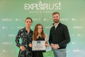 Trzy osoby pozujące do zdjęcia na tle zielonej ścianki z powtarzającymi się logotypami. W tle u góry duży napis: „EXPLORUS!” oraz poniżej „NAUKA BEZ GRANIC”. Na ściance widać też mniejsze loga „EXPLORUS! NAUKA BEZ GRANIC” i „Minister Edukacji” z godłem. Pośrodku stoi dziewczynka o jasnej cerze i długich, falowanych, jasnobrązowych włosach. Trzyma dyplom. Ma białą bluzkę z krótkimi, bufiastymi rękawami i ciemną sukienkę. Po lewej stoi kobieta o jasnej cerze, z włosami upiętymi do tyłu, w ciemnej sukience w kwiatowy wzór. Po prawej stoi mężczyzna o jasnej cerze, z krótkimi jasnobrązowymi włosami i brodą, w ciemnym swetrze na jasnej koszuli i w jasnych dżinsach. Trzyma zieloną torbę prezentową z białym napisem „EXPLORUS!”.