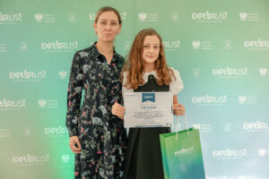Dwie osoby stojące obok siebie na tle zielonej ścianki z logotypami. Po lewej stoi dorosła kobieta o jasnej skórze, z włosami w kolorze ciemny blond/brąz, związanymi do tyłu. Ma na sobie ciemną, długą sukienkę w kwiatowy wzór i patrzy prosto w obiektyw z neutralnym wyrazem twarzy. Po prawej stoi dziewczynka/nastolatka o jasnej skórze, z długimi, falowanymi włosami w kolorze jasny brąz. Ma na sobie białą bluzkę z krótkimi, bufkami rękawami oraz ciemną sukienkę/pinaforek. Trzyma dyplom i ma lekki uśmiech. Obok dyplomu widać też torbę prezentową w zielono-niebieskim gradiencie z białym napisem „EXPLORUS!”.