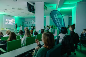 Sala wykładowa z publicznością podczas prezentacji, oświetlona zielonym światłem. W pomieszczeniu stoją rzędy stołów i krzeseł, na których siedzi kilkadziesiąt osób zwróconych w stronę przodu sali. Z przodu, przy ekranie projekcyjnym, stoi kobieta w ciemnej sukience i prezentuje slajd; obok jest mównica, przy której siedzi osoba prowadząca lub obsługująca prezentację. Na ekranie widać kilka czarno-białych grafik i zdjęć twarzy oraz postaci, bez czytelnego tekstu. Po prawej stronie sali stoi wysoki, pionowy baner typu „łezka” z białym napisem.