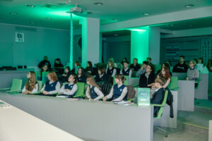 Sala wykładowa z siedzącą publicznością, oświetlona zielonym światłem. Widać kilkadziesiąt osób, głównie nastolatków i młodych dorosłych, siedzących w rzędach przy długich stołach z zielonymi krzesłami; wiele osób ma szkolne stroje: białe koszule i granatowe kamizelki z emblematem. Większość twarzy jest zwrócona w lewą stronę, jakby w kierunku prowadzącego lub ekranu. Na stołach leżą zeszyty i długopisy, a przy kilku miejscach stoją zielone torby reklamowe z białym napisem; na jednej z nich da się odczytać „extratus”. W tle są białe filary, sufit z lampami i projektor zamontowany pod sufitem.