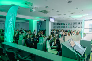 Duża sala wykładowa z publicznością siedzącą w rzędach foteli. Dominuje zielone oświetlenie, które barwi wnętrze i ludzi. Po lewej stronie stoi wysoki, turkusowo-zielony baner w kształcie pióropusza z napisem „EXPLORUJ!” oraz mniejszym tekstem „NAUKA BEZ GRANIC”. W głębi widać kolejny baner z czytelnym napisem „UNIWERSYTET SZCZECIŃSKI”. Na środku i po prawej siedzi kilkadziesiąt osób, głównie młodych; większość ma jasną skórę. Wiele osób patrzy w stronę przodu sali. Na ścianie z tyłu znajduje się rząd czarno-białych portretów w ramkach. Z przodu po prawej jest mównica lub stanowisko prowadzącego; na blacie stoi otwarty laptop z widocznym slajdem na ekranie. Nad salą wisi projektor i widać liczne lampy oraz kratki wentylacyjne w suficie.