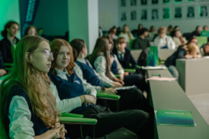 Sala wykładowa wypełniona uczniami siedzącymi w rzędach na zielonych krzesłach. Na pierwszym planie po lewej stronie znajduje się nastolatka o jasnej cerze, długich blond włosach i okularach, ubrana w białą koszulę i granatową kamizelkę; siedzi bokiem z dłońmi splecionymi na kolanach i ma neutralny wyraz twarzy. Obok niej siedzi druga nastolatka o jasnej cerze i rudych, falowanych włosach w podobnym stroju, zwrócona bardziej w stronę aparatu. W dalszych rzędach widać kolejne osoby w szkolnych ubraniach; część rozmawia, część patrzy do przodu. Po prawej stronie na ławce leży zielony folder lub zeszyt z białym napisem, ale tekst jest nieczytelny. W tle na ścianie widać rząd małych portretowych zdjęć. Oświetlenie ma zielonkawy odcień.