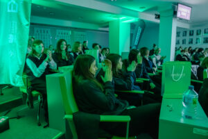 Sala z publicznością siedzącą w rzędach foteli, oświetlona intensywnym zielonym światłem. Wiele osób, głównie młodzież, bije brawo; część ma na sobie ciemne bluzy lub swetry i jasne koszule. Po lewej stronie stoi pionowy zielony baner z napisem „Ministerstwo Edukacji” oraz widocznym godłem. W górnej prawej części jest ekran z niewyraźnym tekstem oraz ściana z szeregiem oprawionych portretów. Na pierwszym planie po prawej na stoliku stoi zielona papierowa torba z napisem „EPLORUJ!” (dalszy drobny tekst nieczytelny) oraz butelka wody; obok leżą metalowe okulary.