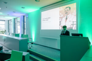Sala konferencyjna z ekranem projekcyjnym i mównicą. W pomieszczeniu dominuje zielone oświetlenie dekoracyjne na ścianach i suficie. Po prawej stronie stoi młoda osoba w ciemnym garniturze, mówiąca do mikrofonu przy mównicy; na mównicy leży otwarty laptop. Na ekranie wyświetlony jest slajd z tekstem „Jan Czochralski (1885–1953)” oraz ilustracją mężczyzny w białym fartuchu laboratoryjnym, w okularach i z wąsem. Po lewej stronie widać długi stół konferencyjny z kilkoma krzesłami, a w tle duże okna.