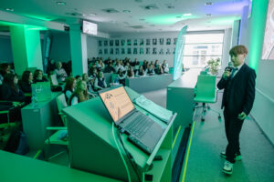 Sala wykładowa z dużą publicznością i chłopcem w garniturze przemawiającym do mikrofonu. W pomieszczeniu dominuje zielonkawe oświetlenie. Po prawej stronie stoi chłopiec o jasnej skórze, w ciemnym garniturze i białej koszuli, trzyma mikrofon w jednej ręce i ma poważny wyraz twarzy. Ma jasnobrązowe włosy z grzywką. Na nogach ma sportowe buty. Na pierwszym planie po lewej jest mównica z otwartym laptopem; na ekranie widać slajd prezentacji, ale tekst jest za mały, żeby go dokładnie odczytać. Obok laptopa leżą kartki i kable. W tle siedzi liczna grupa osób na krzesłach i w ławkach, zwrócona w stronę mówcy. Na ścianie z tyłu widać wiele czarno-białych portretów w ramkach ułożonych w siatkę. Przy oknie stoi wysoka roślina w donicy, a obok są pionowe banery/flagowe standy z logo lub napisem, którego nie da się tu wyraźnie odczytać.