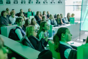 Sala wykładowa z publicznością siedzącą w kilku rzędach ławek, oświetlona silnym zielonkawym światłem. Na pierwszym planie siedzi kilka kobiet; jedna z nich, w okularach i z jasnymi włosami, trzyma mały aparat fotograficzny skierowany przed siebie, jakby robiła zdjęcie lub nagrywała. Wokół niej siedzą młodsze osoby, część w szkolnych strojach (białe koszule, ciemne kamizelki), zwrócone w tę samą stronę, w stronę niewidocznego prelegenta lub ekranu. W tle widać więcej osób na wyższych rzędach oraz rząd czarno-białych portretów na ścianie. Po prawej stronie jest duże okno lub przeszklona ściana, wpuszczająca jasne światło.
