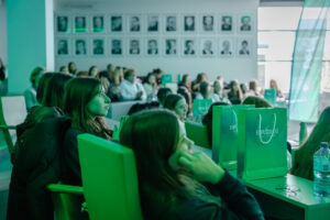 Sala wykładowa z dużą grupą osób siedzących w rzędach i słuchających prezentacji. Dominują młode kobiety; większość ma długie włosy, ubrania w ciemnych kolorach. Na pierwszym planie po prawej stronie stoją na blacie dwa zielone papierowe torby z białymi uchwytami i białym napisem, częściowo uciętym i nie w pełni czytelnym. Obok leży para okularów i stoi przezroczysta butelka wody. W tle na ścianie wisi rząd czarno-białych portretów w ramkach, a z prawej strony widać duże okna wpuszczające jasne światło. Całe zdjęcie ma silny zielonkawy filtr lub oświetlenie.