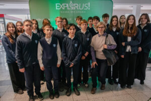 Grupa nastolatków i jedna dorosła kobieta pozująca do zdjęcia na tle zielonej ścianki z napisem „EXPLORUS!” oraz „NAUKA BEZ GRANIC”. To wygląda na zdjęcie szkolne zrobione w budynku. Większość osób ma granatowe stroje szkolne: bluzy lub swetry z białymi koszulami i małe logo na piersi. Dzieci stoją w dwóch rzędach, twarzami do aparatu, z neutralnymi lub lekko uśmiechniętymi wyrazami twarzy. Dorosła kobieta stoi z przodu po prawej stronie; ma krótkie jasne włosy, okulary, jasny sweter na koszuli we wzór i czarną torebkę na ramieniu. W tle po lewej widać przeszklone ściany i fragment wnętrza.
