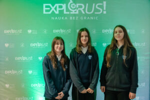 Zielone tło fotograficzne z dużym białym napisem „EXPLORUS!” oraz hasłem „NAUKA BEZ GRANIC”. Na tle stoją trzy nastolatki o jasnej karnacji w ciemnych, szkolnych bluzach; wszystkie mają długie, brązowe włosy. Dwie osoby (po lewej i pośrodku) stoją z dłońmi złożonymi z przodu i lekko się uśmiechają. Osoba po prawej stoi bardziej wyprostowana, ma neutralny wyraz twarzy, a na bluzie widać okulary wiszące przy dekolcie. Na tle powtarzają się mniejsze logotypy „EXPLORUS!” oraz napisy „Minister Edukacji”