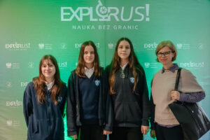Grupa czterech osób stojących przed zieloną ścianką eventową z dużym napisem „EXPLORUS!” i hasłem „NAUKA BEZ GRANIC”. W tle powtarzają się mniejsze logotypy „EXPLORUS!”, godło Polski oraz napis „Minister Edukacji”. Od lewej stoją trzy nastolatki w ciemnych szkolnych bluzach z tarczą-herbem na piersi; dwie mają długie brązowe włosy, a jedna (najwyższa) ma bardzo długie włosy i ma okulary zawieszone na szyi. Po prawej stoi dorosła kobieta o jasnej cerze, z krótkimi blond włosami i okularami, w jasnym swetrze na koszuli w drobny wzór, z czarną torbą na ramieniu. Wszystkie osoby są zwrócone przodem do aparatu i mają neutralny, lekko uśmiechnięty wyraz twarzy.