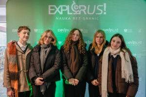 Grupa pięciu nastolatków stojących w rzędzie na tle zielonej ścianki promocyjnej z logotypami. W tle u góry jest duży biały napis „EXPLORUS!” oraz mniejszy napis „NAUKA BEZ GRANIC”. Na ściance w powtarzającym się wzorze widać też mniejsze logotypy „EXPLORUS!”, napis „Minister Edukacji” oraz godło Polski.