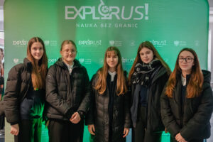 Pięć młodych kobiet stojących w rzędzie na tle zielonej ścianki promocyjnej z napisem „EXPLORUS! NAUKA BEZ GRANIC”. Wszystkie mają jasną karnację i długie włosy, ubrane są głównie w ciemne kurtki zimowe. Druga od lewej ma okulary. Czwarta od lewej ma gruby, ciemny szalik w jasną kratę. Na ściance w tle powtarzają się mniejsze logotypy „EXPLORUS!” oraz „Minister Edukacji” z godłem.