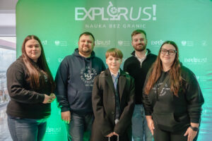 Grupowe zdjęcie pięciu osób stojących przed zieloną ścianką promocyjną. W tle na ściance widoczny jest duży napis „EXPLORUS!” oraz poniżej „NAUKA BEZ GRANIC”. Powtarzają się też mniejsze logotypy i napisy, w tym „Minister Edukacji” oraz „EXPLORUS! NAUKA BEZ GRANIC”. Od lewej stoi młoda kobieta z długimi, prostymi brązowymi włosami, w czarnej bluzie i dżinsach. Obok stoi mężczyzna w granatowej bluzie z kapturem z białym nadrukiem „UNIWERSYTET SZCZECIŃSKI” (z datą „EST. 1985”), ma krótko przycięte włosy i zarost. Na środku stoi chłopiec w ciemnym płaszczu, białej koszuli i krawacie, z plecakiem, ma jasnobrązowe włosy z grzywką. Po jego prawej stronie stoi mężczyzna z krótkimi jasnobrązowymi włosami i brodą, w ciemnym swetrze na jasnej koszuli. Skrajnie po prawej stoi kobieta w okularach, z długimi brązowymi włosami, w czarnej bluzie z podobnym nadrukiem „UNIWERSYTET SZCZECIŃSKI” i czarnych spodniach. Oświetlenie ma zielonkawy odcień, szczególnie na ubraniach i tle.