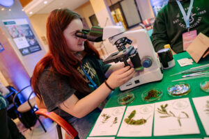 Młoda kobieta z długimi rudymi włosami siedząca przy stole i patrząca przez okular mikroskopu laboratoryjnego. Ma jasną cerę i trzyma jedną ręką pokrętło regulacji mikroskopu, skupiona na obserwacji preparatu na stoliku. Na stole leży zielona mata, kilka pęset i metalowych narzędzi, małe szalki Petriego z fragmentami roślin lub mchu oraz trzy karty z ilustracjami i opisami roślin (tekst na kartach jest zbyt mały, żeby go dokładnie odczytać). Z prawej strony częściowo widać inną osobę w zielonej bluzie z identyfikatorem na smyczy. Tło wygląda jak wnętrze budynku z plakatami i oświetleniem sufitowym, sugerujące stoisko edukacyjne lub pokaz.