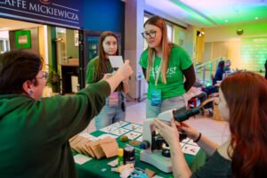 Stoisko edukacyjne z mikroskopem na stole, przy którym kilka młodych osób rozmawia i prezentuje próbki. W centrum stoi młoda kobieta w zielonej koszulce z identyfikatorem na smyczy; ma długie proste włosy i okulary. Obok niej stoi druga młoda kobieta w podobnej zielonej koszulce i z identyfikatorem, trzyma notatnik i długopis. Po lewej stronie kadru osoba w zielonej bluzie trzyma w górze małą kartkę. Po prawej stronie młoda kobieta o rudych włosach obsługuje biały mikroskop ustawiony na stole. Na stole z zielonym obrusem leżą papierowe koperty, nożyczki, klej w sztyfcie, mała puszka oraz kilka kart z rysunkami lub opisami roślin i drobnymi okazami/próbkami. W tle widać wejście do lokalu z napisem „CAFFE MICKIEWICZA” oraz korytarz z zielonym oświetleniem i siedzącymi ludźmi.