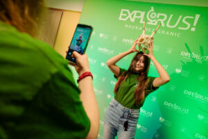 Młoda kobieta pozująca do zdjęcia na tle zielonej ścianki promocyjnej „EXPLORUS! NAUKA BEZ GRANIC”. Stoi przodem i uśmiecha się, ma długie brązowe włosy i jasną cerę. Jest ubrana w zieloną koszulkę i jasnoniebieskie jeansy; na wysokości rozporka zwisa czarny element (wygląda jak smycz lub klucze). Nad głową trzyma biało-beżową czaszkę zwierzęcia z rogami. Po lewej stronie kadru inna osoba fotografuje ją smartfonem; na ekranie telefonu widać tę samą pozę. Na ściance w tle powtarzają się logotypy i napisy, m.in. „EXPLORUS!”, „NAUKA BEZ GRANIC” oraz „Minister Edukacji”.