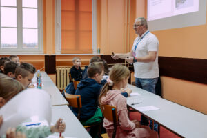 Sala lekcyjna z prowadzącym stojącym przy ławkach i grupą dzieci siedzących i piszących. Po prawej stronie stoi mężczyzna w średnim wieku, o jasnej skórze, krótkich siwych włosach i okularach. Ma na sobie białą koszulkę polo, dżinsy oraz identyfikator na niebieskiej smyczy; w dłoniach trzyma mały czarny pilot lub marker i gestykuluje, jak podczas tłumaczenia. Po lewej i na środku siedzi kilkanaścioro dzieci w wieku szkolnym przy długich szarych ławkach, na drewnianych krzesłach. Większość ma przed sobą kartki i długopisy; część dzieci pochyla się nad zadaniami, a jedno dziecko w czarnej bluzie patrzy w stronę prowadzącego. Na stole stoi wysoka butelka z kolorową etykietą. W tle są duże okna z pomarańczowymi roletami, kaloryfer pod oknem i pomarańczowe ściany z ciemnym pasem. W prawym górnym rogu widać fragment rzutowanego slajdu, ale tekst jest nieczytelny.