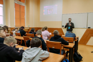 Sala lekcyjna z uczniami siedzącymi w ławkach i prowadzącym stojącym z przodu przy tablicy. Widać kilkanaście osób, głównie dzieci i nastolatków, większość odwrócona tyłem i pochylona nad zeszytami, jakby notowała lub pisała. Z przodu po prawej stoi dorosły mężczyzna z krótkimi włosami i brodą, w ciemnym swetrze i dżinsach, z identyfikatorem na smyczy; ma uniesione dłonie w geście tłumaczenia. Na ścianie za nim jest biała tablica oraz rzutnik wyświetlający slajd, ale tekst na slajdzie jest nieczytelny. Po lewej stronie są wysokie okna z pomarańczowymi roletami.