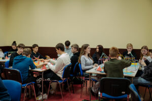 Grupa nastolatków siedzących przy stołach w sali, pracujących nad kolorowymi modelami geometrycznymi ze słomek i łączników. Widać kilka stołów ustawionych w rzędzie, a na blatach leżą patyczki/słomki w różnych kolorach, trójkątne elementy z papieru lub kartonu, nożyczki i inne drobne przybory. Uczniowie są w codziennych ubraniach (bluzy, swetry), skupieni na składaniu konstrukcji; część rozmawia, część patrzy na modele. Tło to jasna, jednolita ściana, a podłoga jest czerwonawa; krzesła są niebiesko-czarne.