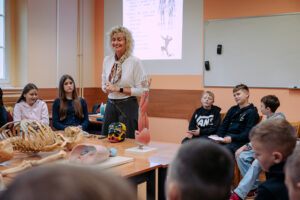 Kobieta prowadząca zajęcia w klasie, stojąca przy stole z pomocami anatomicznymi. Ma jasną skórę i kręcone blond włosy do ramion, białą bluzkę, wzorzysty szalik przy szyi, ciemne spodnie i zegarek na nadgarstku; uśmiecha się i ma dłonie złożone przed sobą. Wokół stołu siedzi grupa nastolatków. Po lewej stronie dwie dziewczyny (obie o jasnej skórze); jedna ma ciemne, długie włosy i jasną bluzę z kapturem, druga ma bardzo długie, ciemne włosy i ciemny sweter. Po prawej stronie kilku chłopców o jasnej skórze w ciemnych bluzach; jeden ma na bluzie duży napis „VANS”. Na stole leżą modele i preparaty anatomiczne: fragment szkieletu (żebra i kości), czaszka, kolorowy model głowy/czaszki, duży model mięśni ramienia i dłoni na podstawce oraz przekrojowy model narządu (prawdopodobnie ucha) na białej podstawie. W tle widać tablicę suchościeralną oraz rzutnik z wyświetlonym slajdem z rysunkami anatomicznymi i tekstem, ale tekst jest zbyt rozmyty, by go wiernie przepisać.