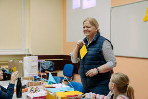 Kobieta stojąca w sali lekcyjnej, trzymająca w dłoni żółtą kartkę i uśmiechająca się. Ma jasną skórę, jasnoblond włosy związane z tyłu, granatową pikowaną kamizelkę na koszuli w drobny wzór oraz zegarek na lewym nadgarstku. Przed nią jest stół zasypany materiałami do zajęć: kolorowe kartki, teczki, nożyczki, taśmy, geometryczne modele z patyczków i rurek, czarny bidon oraz pudełko z napisem „ColorChoice”. Wokół stołu siedzą dzieci; jedno po lewej stronie podnosi żółtą kartkę, a drugie po prawej ma włosy związane w kucyk i sweter w różowo-białe pasy. W tle są niebieskie krzesła, torba i biała tablica na ścianie z żółtą kartką przypiętą w prawym górnym rogu.