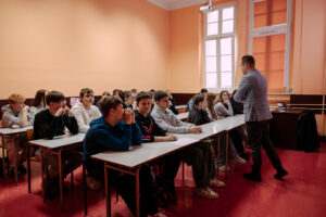 Sala lekcyjna z grupą nastolatków siedzących w ławkach i mężczyzną stojącym z przodu po prawej stronie. Uczniowie siedzą w kilku rzędach przy długich stołach, większość ma swobodne ubrania (bluzy, swetry), część opiera głowy na dłoniach lub ma ręce złożone na stole. Mężczyzna stoi bokiem do grupy, ubrany w jasnoszarą marynarkę i ciemne spodnie, wygląda jak prowadzący zajęcia. Pomieszczenie ma jasnopomarańczowe ściany, czerwono-bordową podłogę i wysokie, białe okna po prawej stronie; przy suficie widać projektor. W tle są dodatkowe ławki i krzesła, a przy ścianie po prawej stoi biurko z otwartym laptopem.