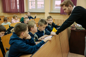 Sala lekcyjna z grupą dzieci siedzących w ławkach oraz dorosłym mężczyzną pochylonym nad pierwszym rzędem. Mężczyzna ma krótkie brązowe włosy i brodę, jest ubrany na ciemno i podaje dziecku żółtą papierową figurę przypominającą origami. Na pierwszym planie kilku chłopców w granatowych bluzach patrzy w stronę nauczyciela lub na swoje zeszyty; na ławce leżą kartki i długopisy. W tle widać kolejne dzieci, drewniane ławki, okna z bordowymi zasłonami oraz grzejnik pod oknami. Po prawej stronie stoi biurko z otwartym laptopem i kablami.