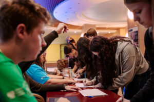 Grupa młodych ludzi przy długim stole w budynku, pochylona nad kartkami i formularzami. Kilka osób siedzi po lewej stronie stołu, a kilka stoi po prawej i podpisuje lub wypełnia dokumenty długopisami. Na pierwszym planie po lewej stronie widać rozmyty profil młodego mężczyzny w zielonej koszulce. Po prawej stronie na pierwszym planie stoi młoda kobieta o długich, kręconych ciemnych włosach w jasnej bluzie z kapturem, pisząca na formularzu. W tle widać wnętrze z ciepłym oświetleniem i okrągłym sufitem.