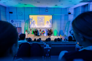 Sala wykładowa z publicznością na pierwszym planie i sceną na dole, oświetlona niebiesko-zielonym światłem. Na scenie stoi grupa kilku osób przy mównicy i długim stole; pośrodku widać kolorowe koło do losowania (jak koło fortuny). Nad nimi jest duży ekran z kodem QR oraz grafiką z dwiema osobami w białych fartuchach.