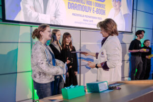 Scena wręczania nagrody na wydarzeniu w sali z dużym ekranem projekcyjnym. Kobieta w jasnej kurtce i dżinsach stoi z lewej strony i odbiera dokument lub dyplom od osoby po prawej w białym fartuchu, która trzyma kilka kartek. Za odbierającą stoją dwie młode kobiety w ciemnych ubraniach, obserwujące sytuację. W tle po prawej widać dwie kolejne osoby w ciemnych strojach, jedna trzyma kolorowe pudełko. Na dole kadru jest stół z kilkoma przedmiotami, w tym zielonym stojakiem/pojemnikiem i innymi akcesoriami.