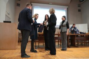 Grupa pięciu dorosłych osób na sali wykładowej podczas wręczania nagrody. Scena wygląda na uroczystość lub konkurs: w centrum dwie kobiety trzymają puchar (złoty kielich) oraz dyplomy lub teczki, a jedna z nich podaje drugiej torbę upominkową. Po lewej stronie stoi mężczyzna w ciemnym garniturze, zwrócony bokiem, trzyma w rękach dokumenty. Obok niego kobieta w długim, ciemnym płaszczu pochyla się w stronę środka i przekazuje torbę. W środku stoi młoda kobieta z długimi ciemnymi włosami, trzyma puchar i papiery. Z przodu, tyłem do obiektywu, stoi kobieta w czarnym stroju z jasnymi włosami upiętymi w kok, również trzyma puchar lub dyplom. Po prawej stoi kobieta w okularach, w szarej kamizelce na czarnym golfie i szarych spodniach, obserwuje sytuację. W tle, przy stole, jest jeszcze jedna kobieta w niebieskiej marynarce, zajmuje się torbami lub materiałami. Pomieszczenie ma drewnianą podłogę, długie stoły i krzesła. Na ścianie z tyłu jest duży ekran projekcyjny z wyświetloną stroną internetową; tekst jest zbyt niewyraźny do dokładnego odczytania. W prawym dolnym rogu zdjęcia widać mały podpis: „fot. Magdalena Seredyńska/Uniwersytet Szczeciński”.