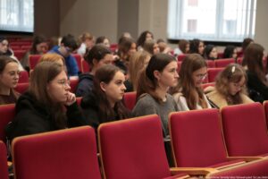 Grupa młodych ludzi siedzących w sali wykładowej na czerwonych, tapicerowanych fotelach ustawionych w rzędach. Większość osób to młode kobiety o jasnej karnacji, z długimi włosami (głównie brązowymi i blond), część w okularach. W pierwszych rzędach kilka osób ma skupione, poważne miny; jedna opiera brodę na dłoni, inna patrzy prosto przed siebie, a po prawej stronie jedna osoba spogląda w dół na otwarty notes lub zeszyt. W tle widać kolejne rzędy siedzeń z większą liczbą uczestników, także kilku młodych mężczyzn. Po prawej stronie znajdują się duże okna z pionowymi żaluzjami, przez które wpada jasne światło dzienne. W prawym dolnym rogu zdjęcia jest mały podpis: „fot. Magdalena Seredyńska/Uniwersytet Szczeciński”.
